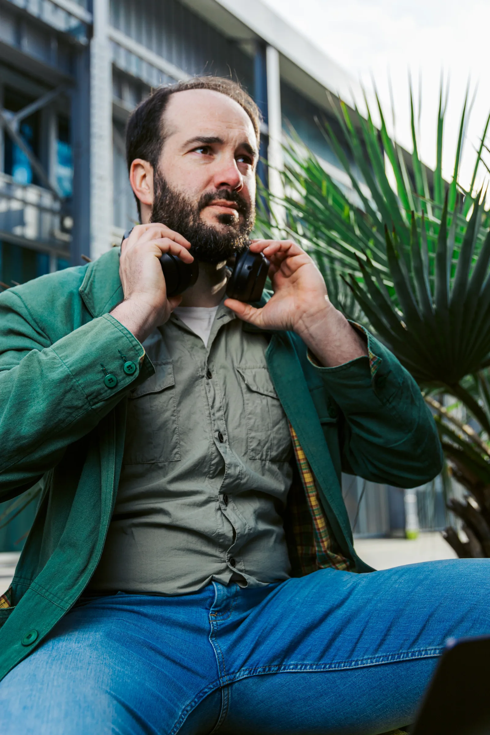 Développeur freelance assis ajustant son casque audio près d'un palmier, en contre-plongée