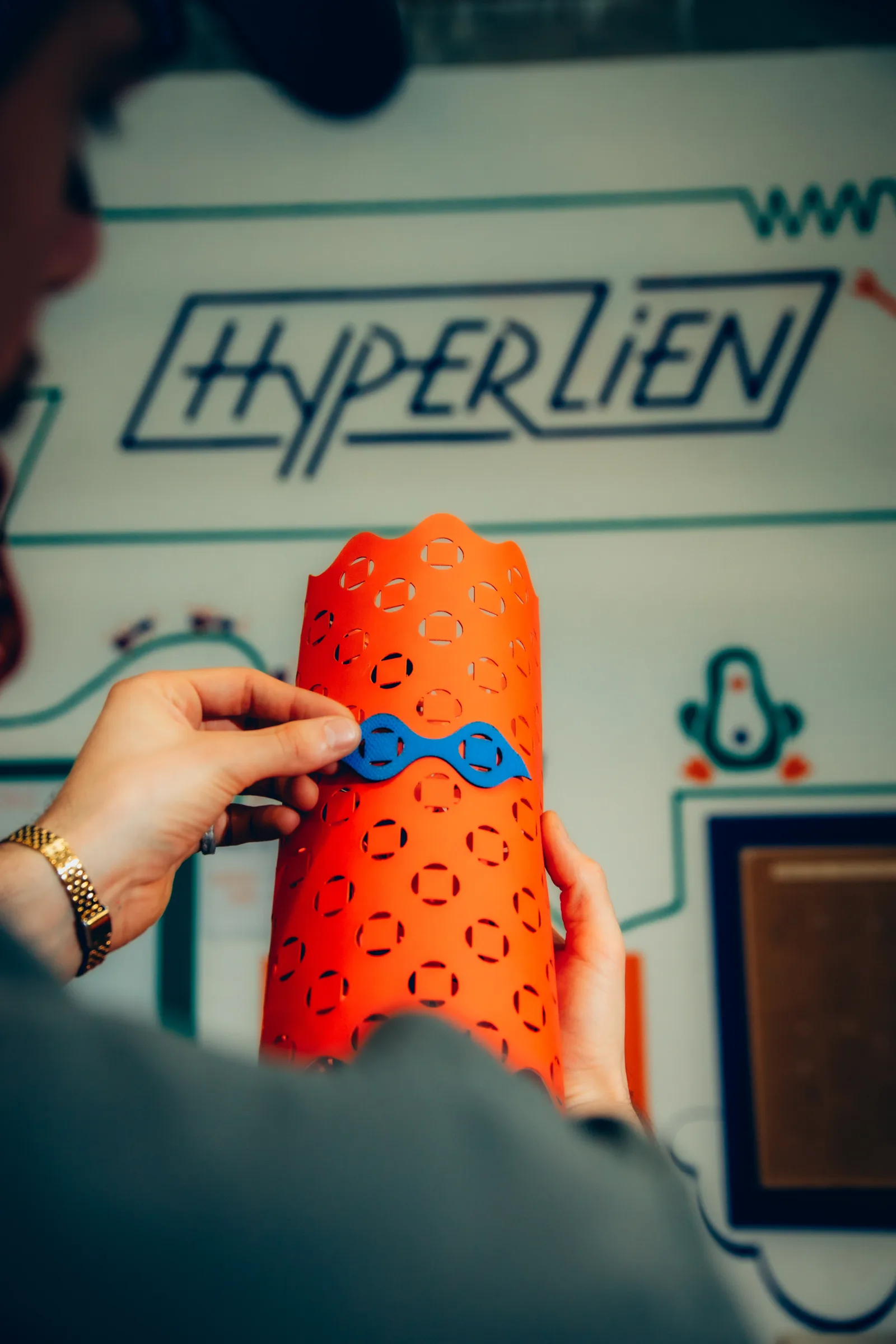 Mains de Loïc assemblant une pièce bleue sur un cylindre Noizy orange au Fablab Hyperlien