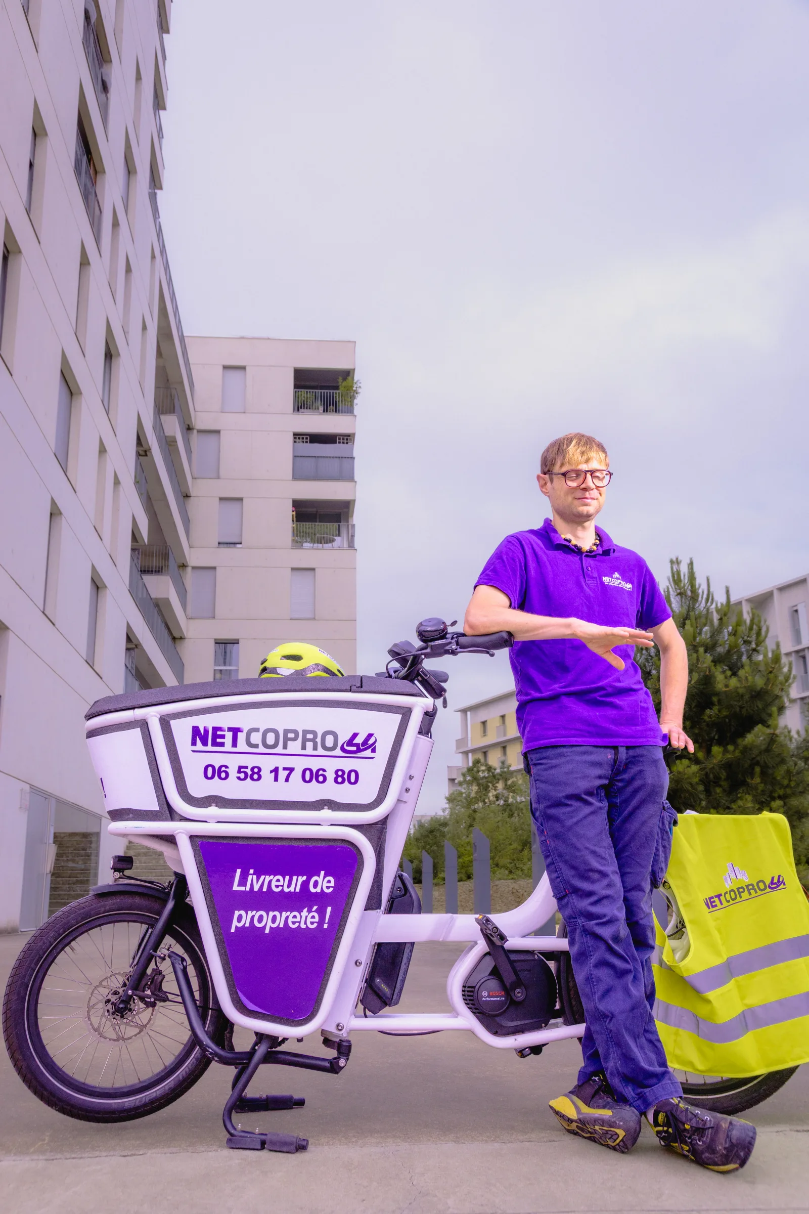Agent Netcopro44 adossé à son vélo cargo violet au pied d'immeubles, en contre-plongée