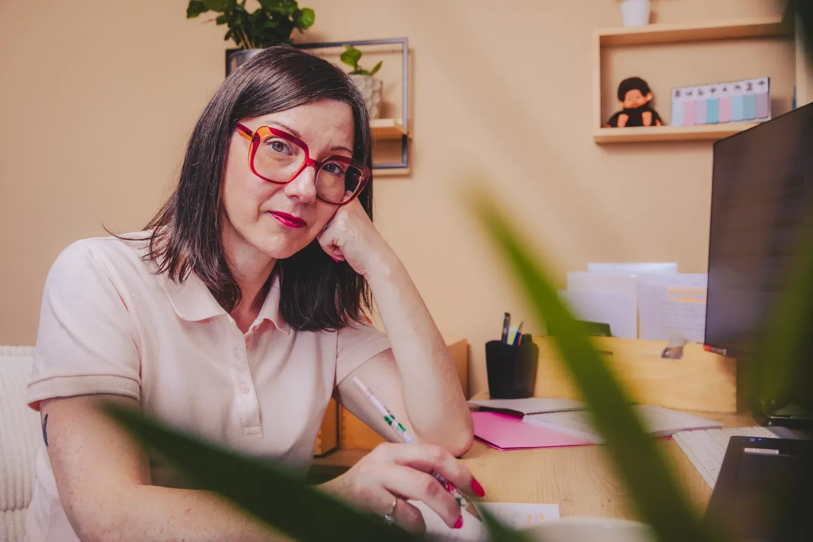 Nelly Audrain, assistante administrative virtuelle, accoudée à son bureau, lunettes rouges, stylo en main, plante floue au premier plan