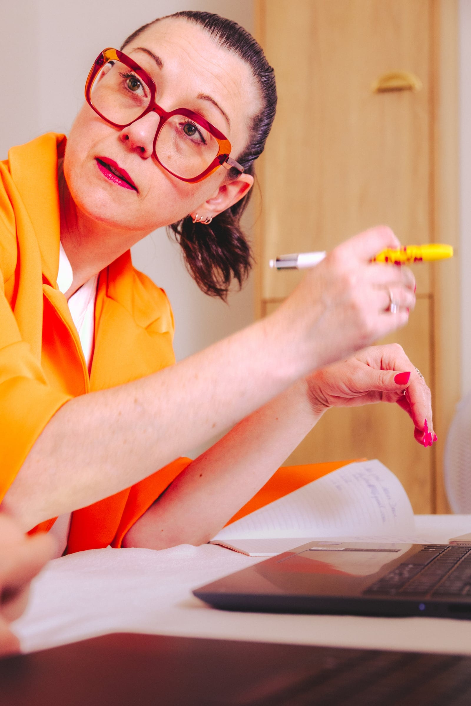 Nelly Audrain en veste orange et lunettes rouges, stylo en main à son bureau